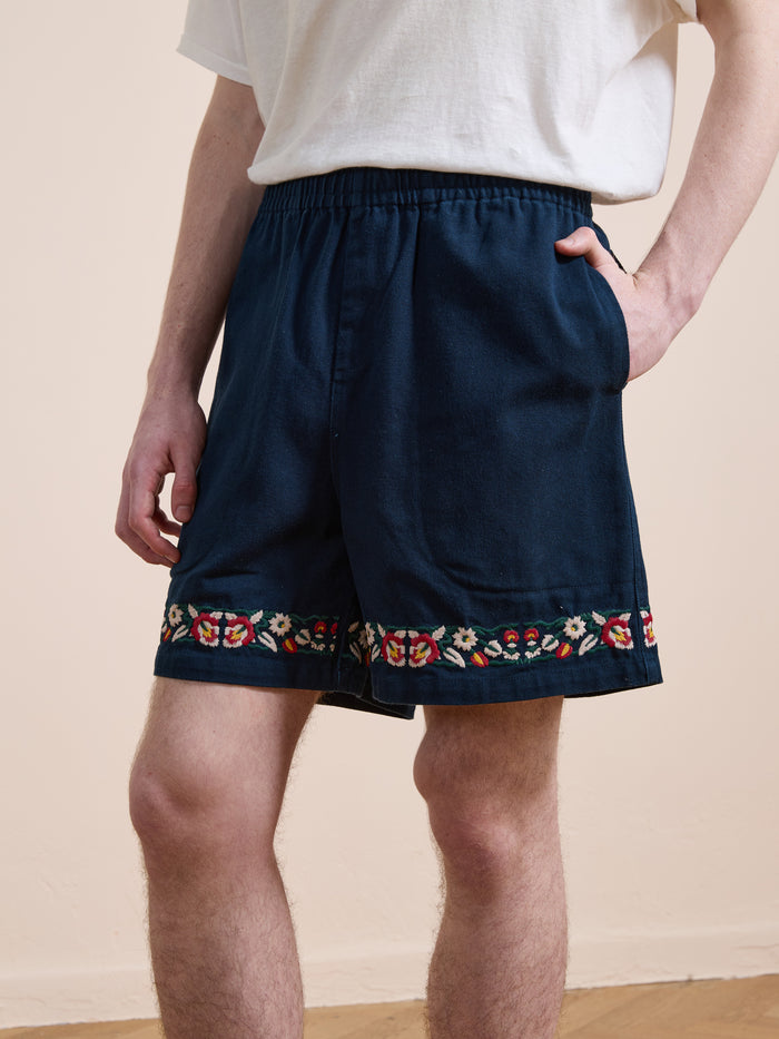 A person wears a white t-shirt and Found Horse Equine Twill Shorts with floral embroidery near the hem, standing indoors with one hand in a pocket.