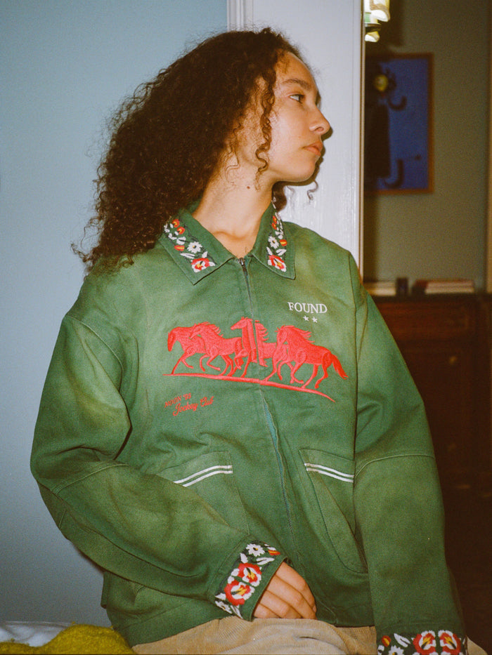 A person with curly hair wears the Found Jockey Club Embroidered Jacket in green, featuring racing horse embroidery and patterned details, indoors in a unisex relaxed fit, looking to the side.