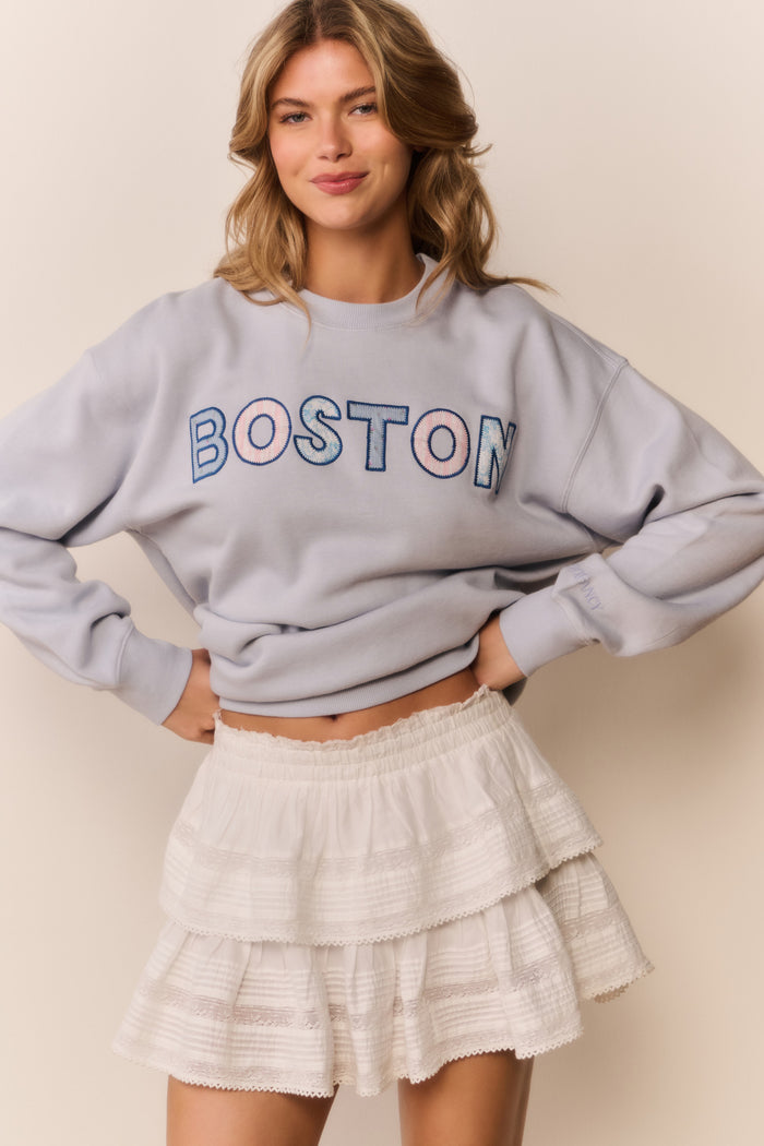 Woman wearing a gray sweatshirt with 'BOSTON' text and a white ruffle skirt on a beige background
