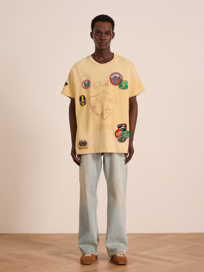 Indoors against a plain wall, a person wears the FOUND Patchwork Camp Tee—unisex, relaxed fit, and finished with vintage-inspired embroidered patches—styled with light blue jeans and brown shoes.