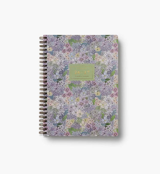 A spiral-bound 2027 daily planner with a dense floral pattern in shades of purple, lavender, and green, featuring a central sage green label with gold text.