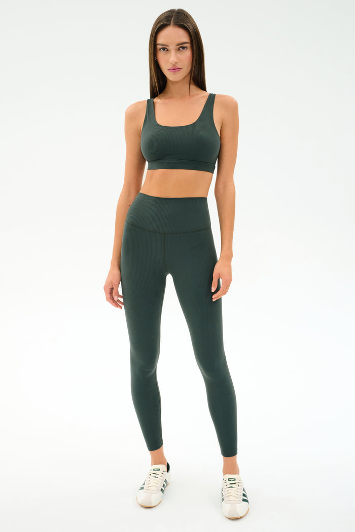 A woman stands against a white background wearing the SPLITS59 Airweight Rib Bra in Military, paired with matching leggings and white sneakers.