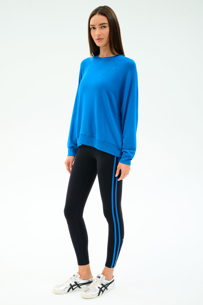 A person wears the SPLITS59 Andie Oversize Fleece Sweatshirt in Classic Blue and black leggings with blue stripes, made from a soft modal spandex blend, paired with white sneakers against a plain white background.