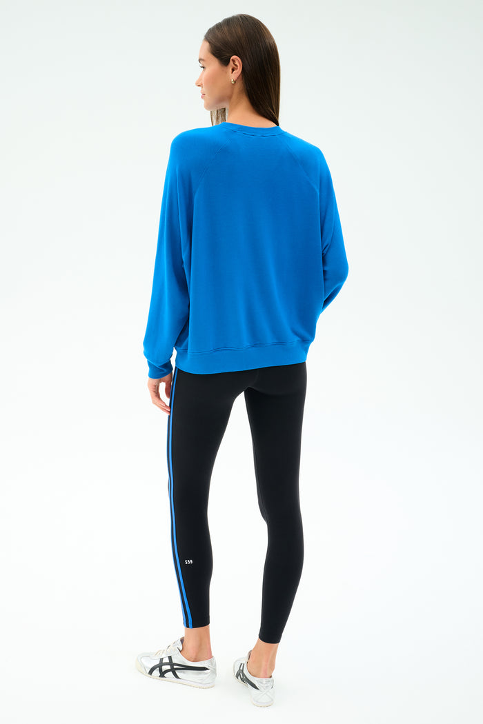 A woman stands facing away, wearing the SPLITS59 Andie Oversize Fleece Sweatshirt in Classic Blue with black leggings featuring a blue stripe, paired with white sneakers against a plain white background.