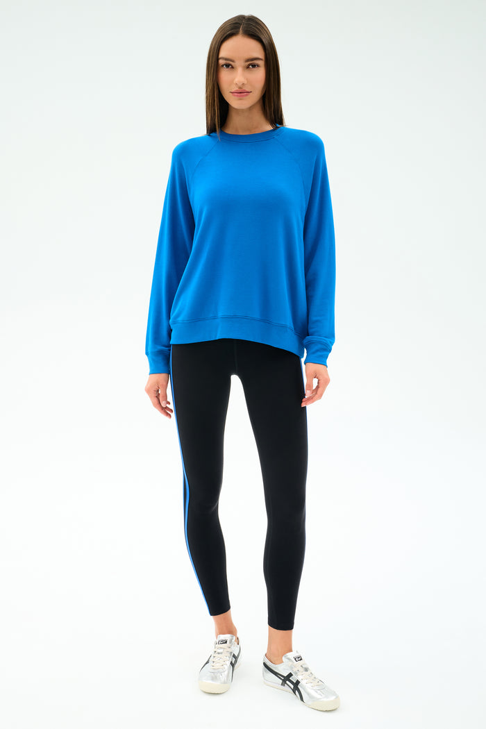 A woman faces forward against a plain light background, wearing the SPLITS59 Andie Oversize Fleece Sweatshirt in Classic Blue and black leggings with a blue stripe, paired with white sneakers.