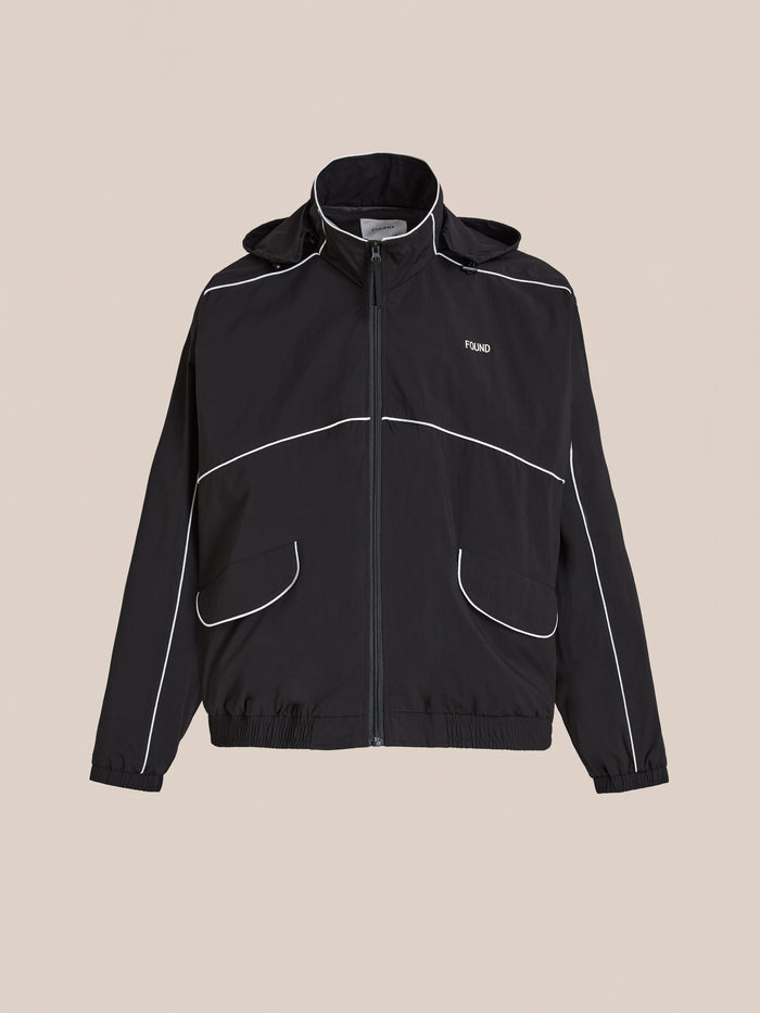 The FOUND Arena Stripe Track Jacket is a unisex black zip-up made from lightweight technical fabric, featuring white piping, two front pockets, elastic cuffs and hem, and a small chest logo. Shown on a beige background.