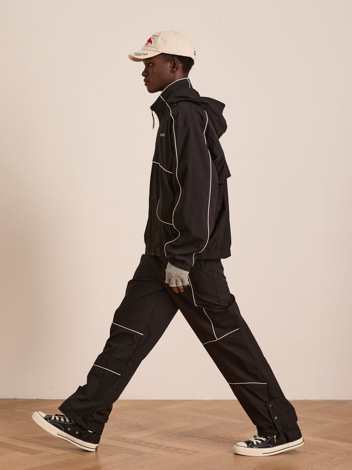 Wearing the FOUND Arena Stripe Track Jacket and matching pants with white piping, a white baseball cap, and black sneakers, a person walks indoors on a wooden floor in style and comfort thanks to the outfit’s lightweight technical fabric.