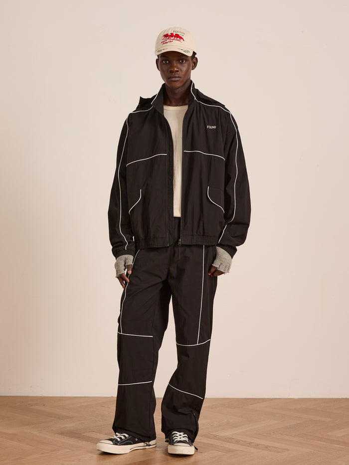 Indoors, a person wears the FOUND Arena Stripe Track Jacket in black with white piping, paired with a beige T-shirt, cap, fingerless gloves, and black lightweight technical sneakers.
