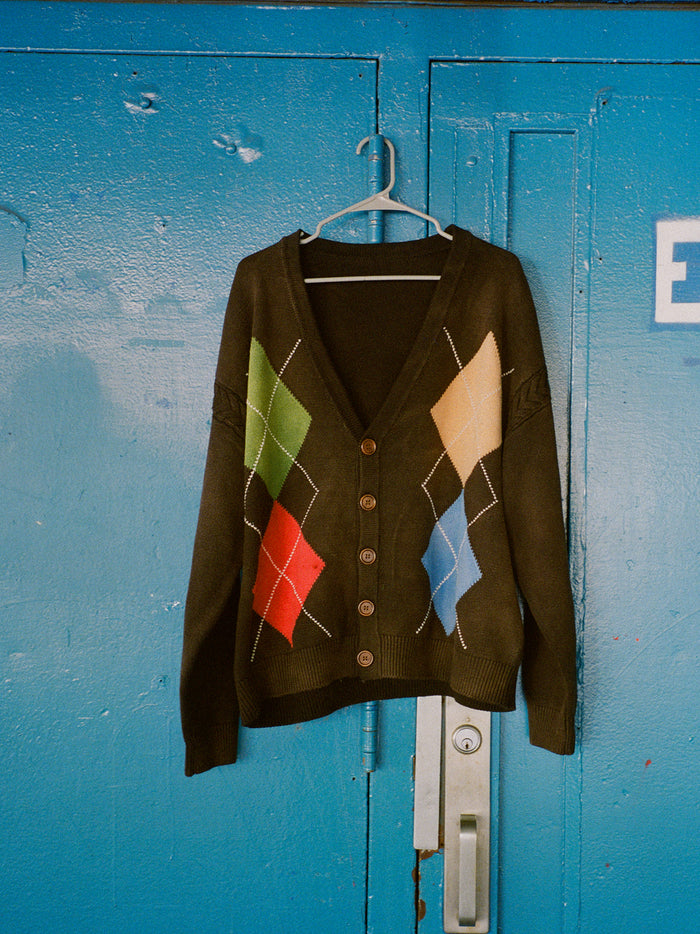 The FOUND Argyle Knit Cardigan, featuring black fabric with red, green, yellow, and blue diamond patterns, hangs on a plastic hanger against a blue metal door.