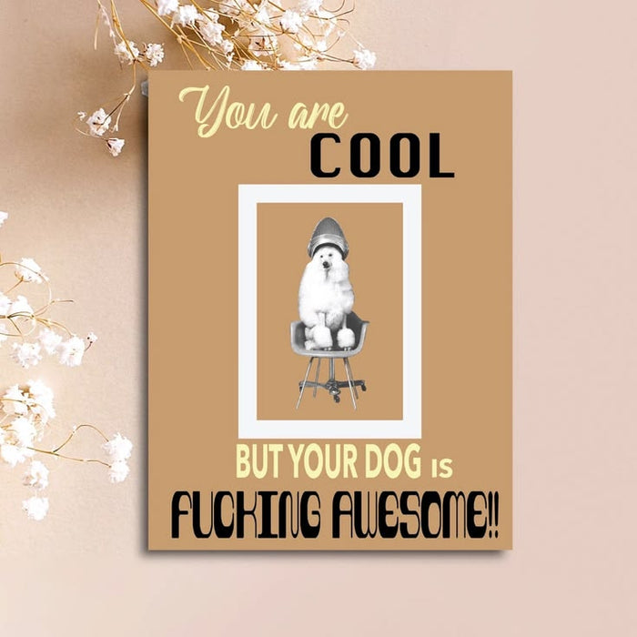 A rectangular printed card or poster with a tan background, featuring the text 'You are COOL BUT YOUR DOG IS FUCKING AWESOME!!' and a central photo of a white dog sitting on a chair. The card is displayed against a beige wall with dried white flowers at the top.