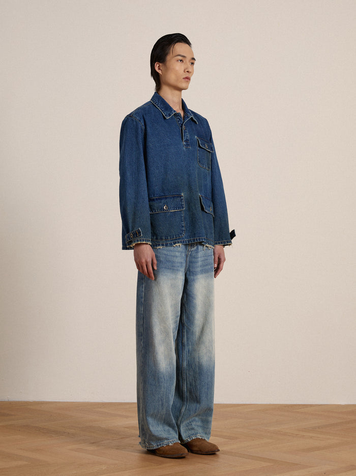 A man stands against a neutral background wearing a Found Azin Denim Pullover Work Shirt and jeans, paired with brown shoes, looking to the side.