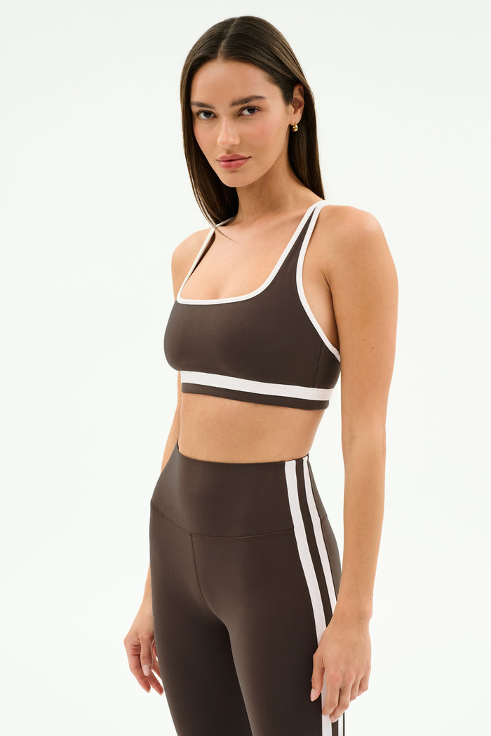 A woman in a SPLITS59 Bianca Rigor Bra - Dark Chocolate/White and coordinating high-waisted leggings with white trim, ideal for gym workouts, stands against a plain white background.