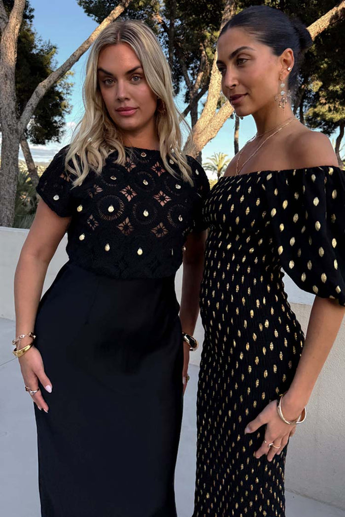 Black Crochet Shell Elodie Dress with gold shell embellishments and a satin skirt, shown on models outdoors.