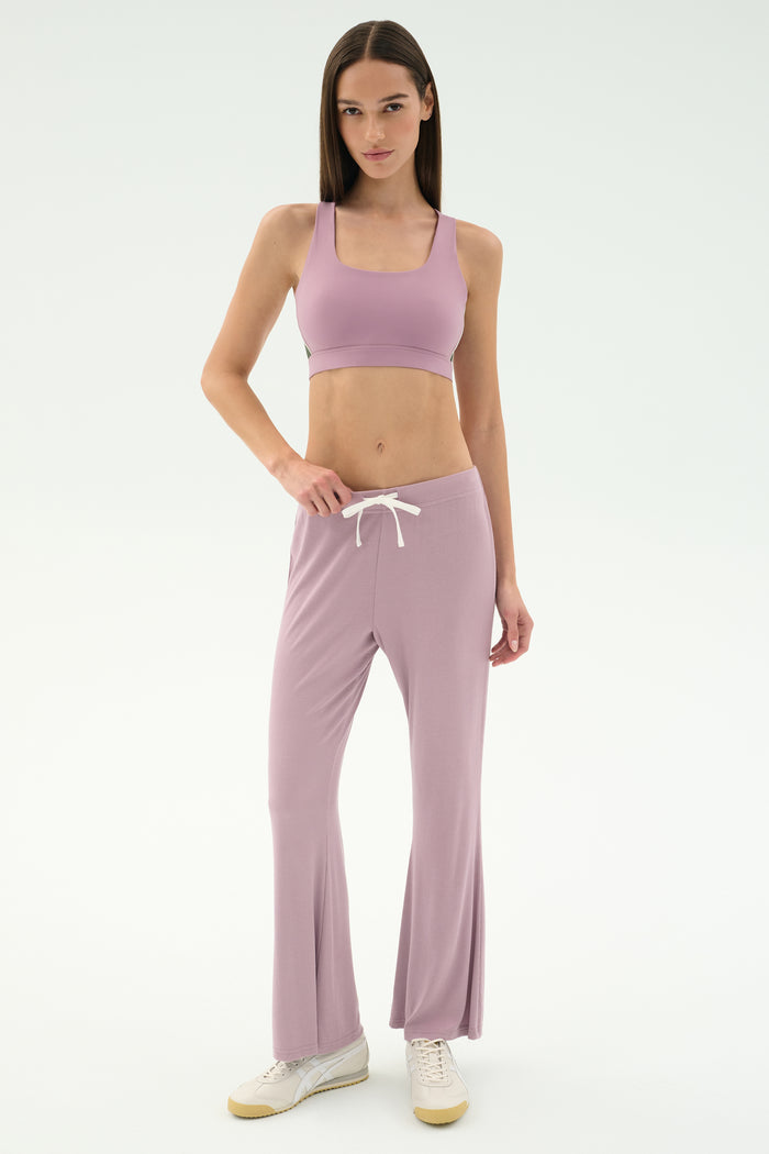 A woman stands against a white background wearing SPLITS59's Blaire Rib Pant in Blush, a coordinating mauve spandex sports bra, and white sneakers.