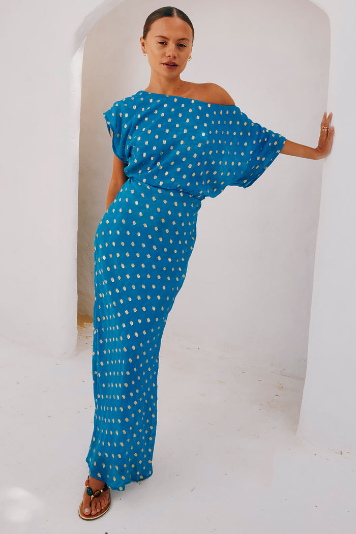 Blue Jacquard Tilly Dress with gold metallic dots and an off-the-shoulder silhouette leaning against a white archway.
