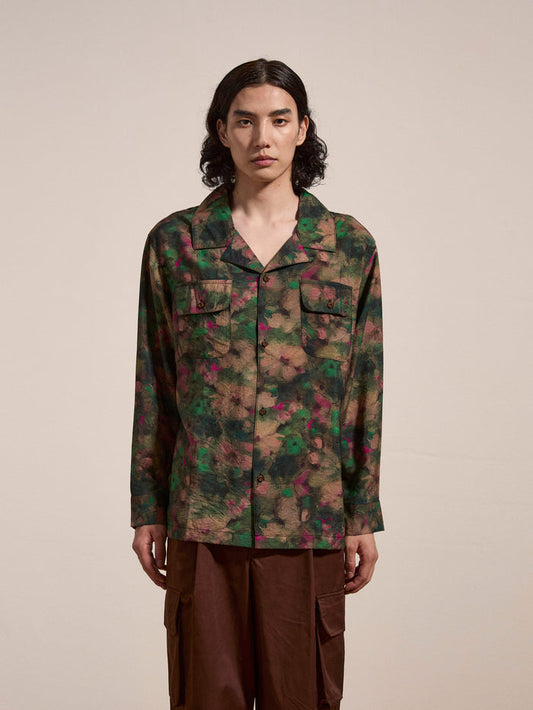 A person with long hair is standing against a plain background, wearing a green and red patterned Charlot LS Camp Shirt from FOUND, featuring dual chest pockets, paired with brown pants.