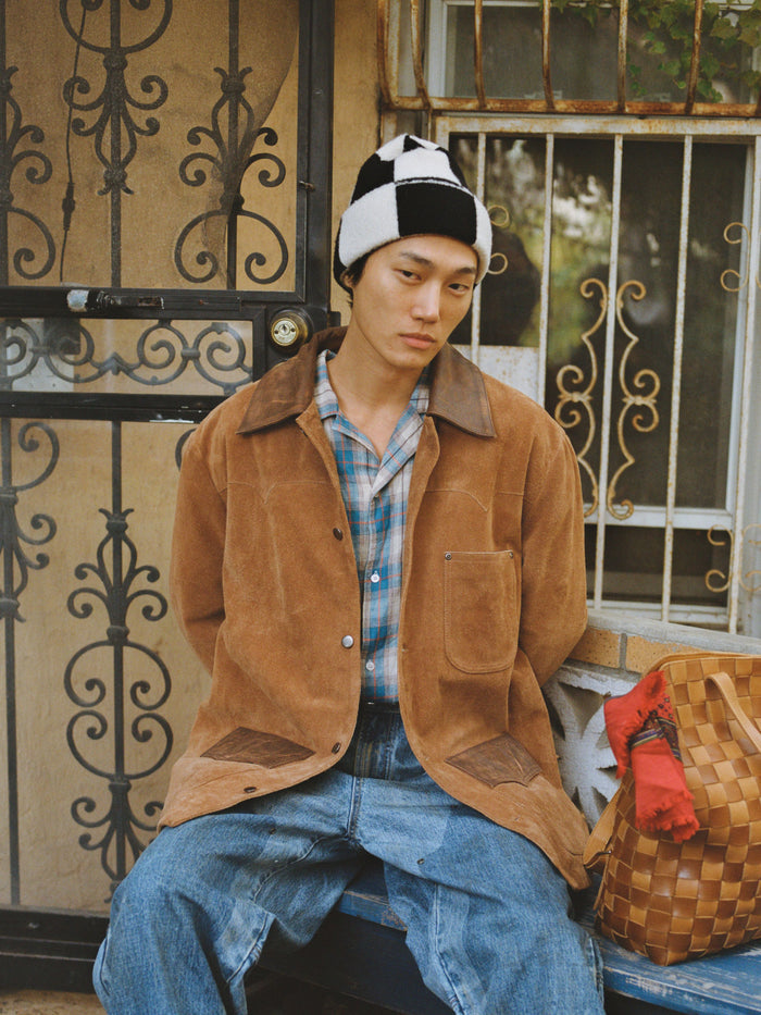 A person wearing a brown jacket, plaid shirt, blue jeans, and the FOUND Check Multi Patch Beanie sits on a bench beside a woven bag, in front of a metal gate and window.