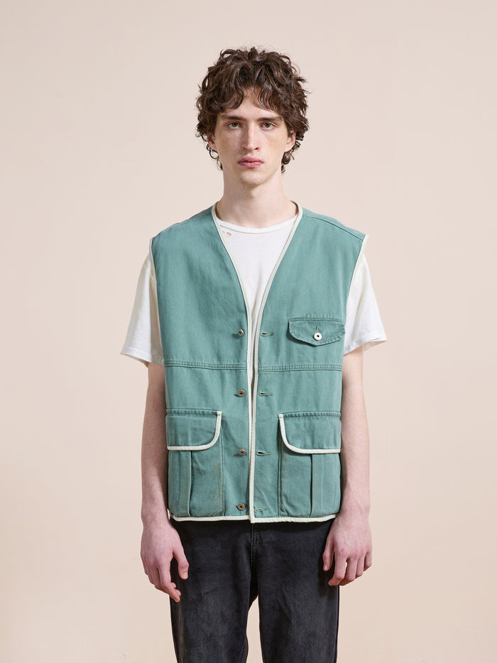 A person stands facing forward wearing the FOUND Cotton Twill Utility Vest in light blue over a white t-shirt and dark pants against a plain beige background.