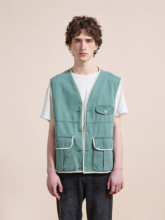 A person stands facing forward wearing the FOUND Cotton Twill Utility Vest in light blue over a white t-shirt and dark pants against a plain beige background.