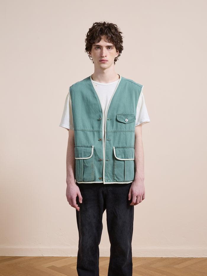 A person stands indoors against a beige wall, wearing a white t-shirt, black pants, and the FOUND Cotton Twill Utility Vest in vintage green with white trim and pockets.