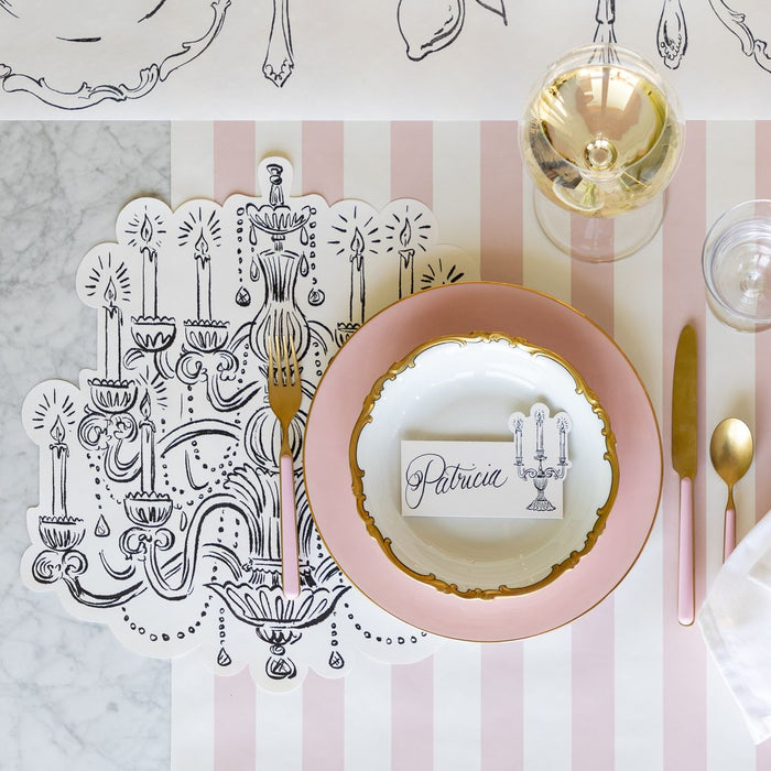 Dinner table setting with pink plates, gold cutlery, and a decorative card on a striped tablecloth.
