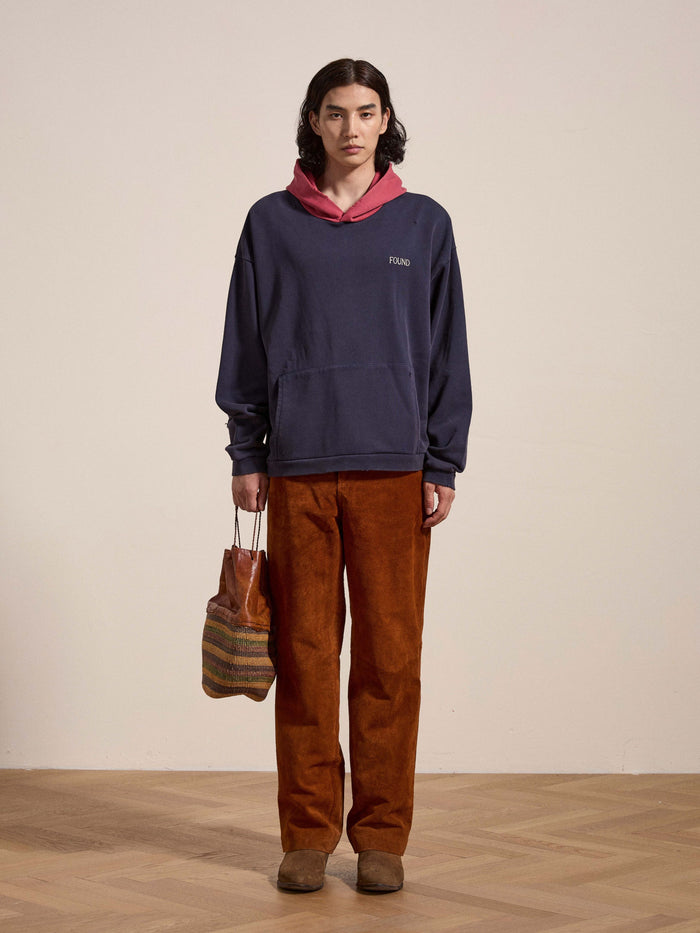A person with shoulder-length hair stands against a plain background, wearing FOUND's blue Distressed Logo Hoodie crafted from French terry cotton, a red hooded layer underneath, brown corduroy pants, and holding a multicolored woven bag. The ensemble exudes a vintage feel.