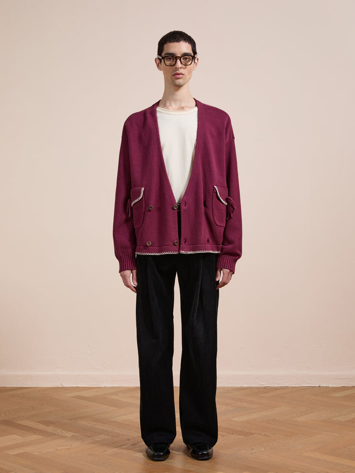 A person stands indoors on a wooden floor, wearing black pants, a white t-shirt, the FOUND Double Breasted Bow Cardigan in maroon, black shoes, and glasses, facing the camera against a plain beige wall.