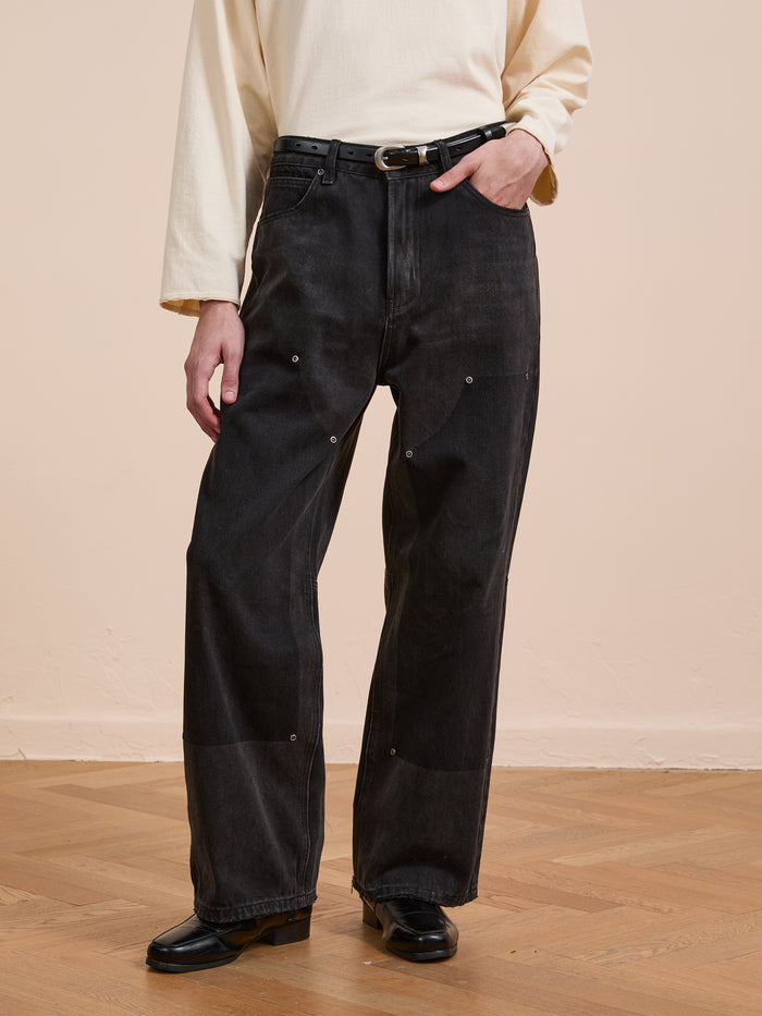 A person wears a long-sleeve cream shirt, FOUND Double Knee Carpenter Jeans in black with decorative stitching and a belt, plus black shoes, standing on a wooden floor against a beige wall.