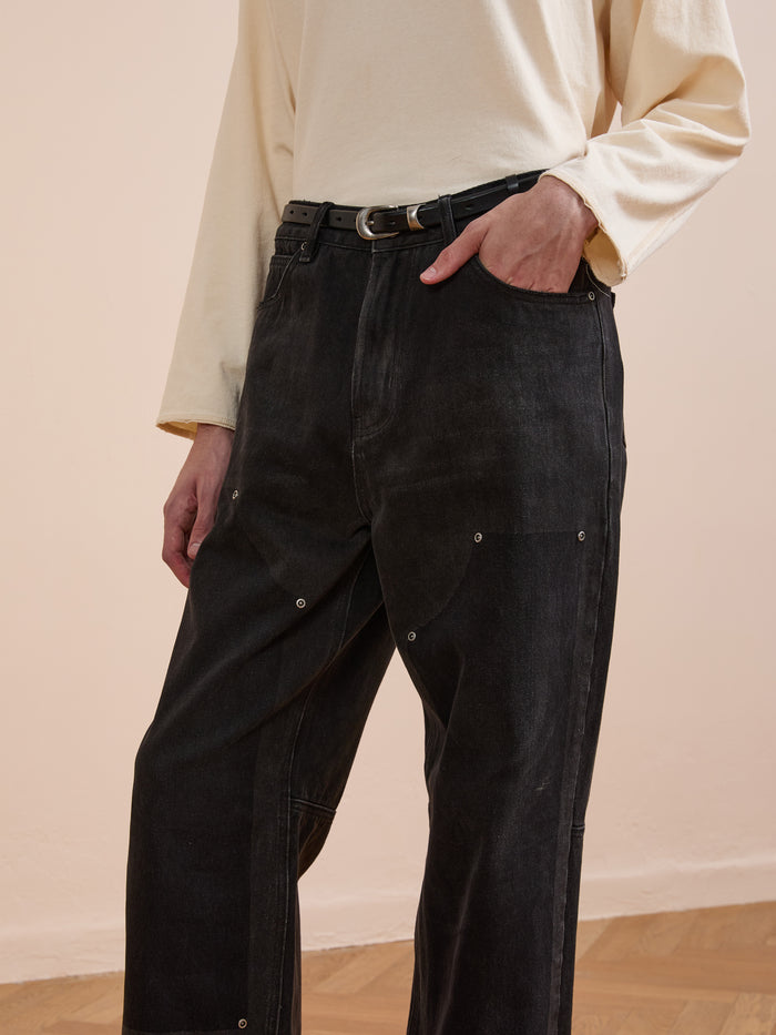 A person wears a long-sleeve beige top tucked into FOUND Double Knee Carpenter Jeans in black, styled with a black belt and standing indoors on a wooden floor.