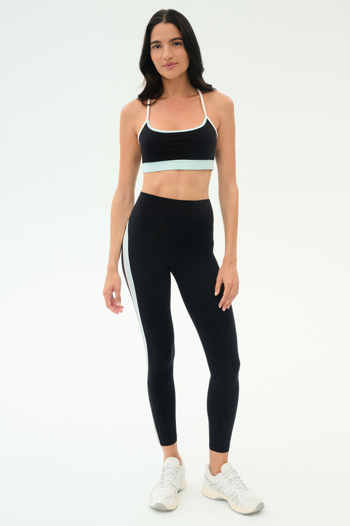 A woman faces forward in a black sports bra, SPLITS59 Emerson Airweight High Waist leggings in Black/Ice with a white side stripe—ideal for hot yoga or Pilates—and white athletic shoes.