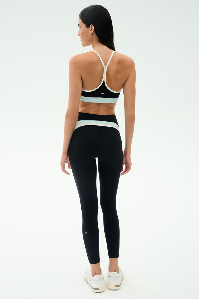 A woman faces away in a black SPLITS59 Emerson Airweight High Waist - Black/Ice legging with white accents and a black sports bra, paired with white sneakers—ideal for Pilates or hot yoga—against a plain white background.