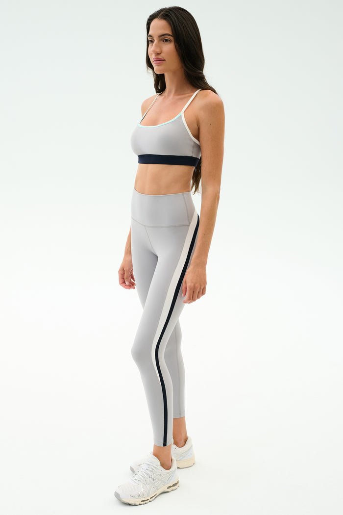 A woman models the SPLITS59 Emerson Airweight High Waist leggings in Dove Grey/Indigo with white sneakers and a light gray sports bra, showcasing a sculpted look ideal for hot yoga against a plain white backdrop.
