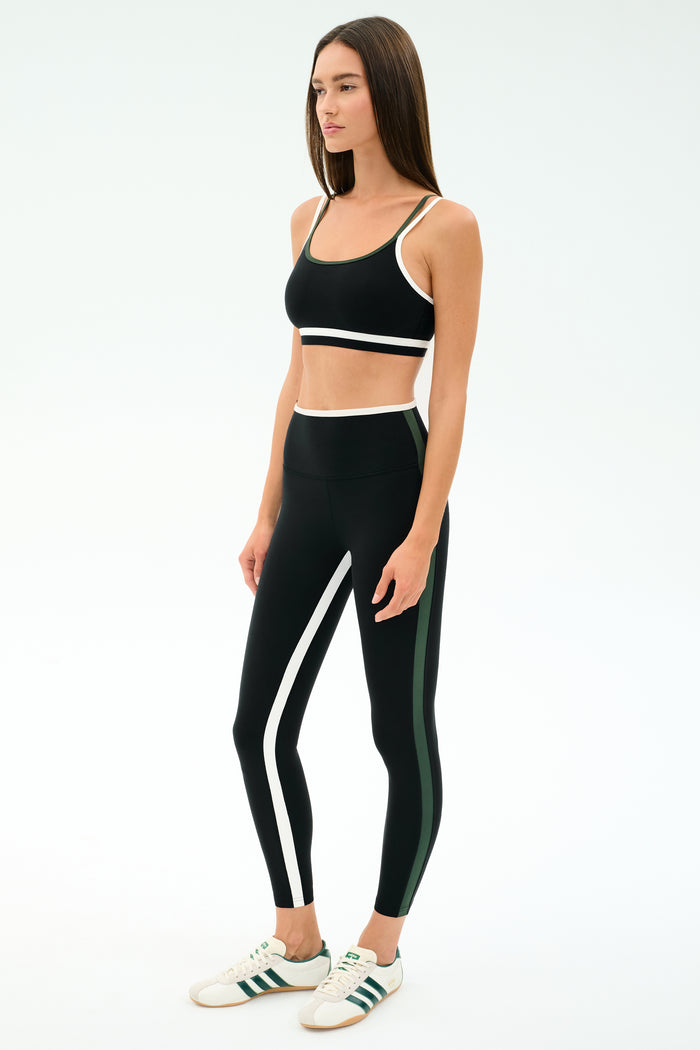 A woman stands in a studio wearing the SPLITS59 Eva Rigor Double Strap Bra in Black/Military, paired with matching black leggings featuring white and green stripes and white sneakers.