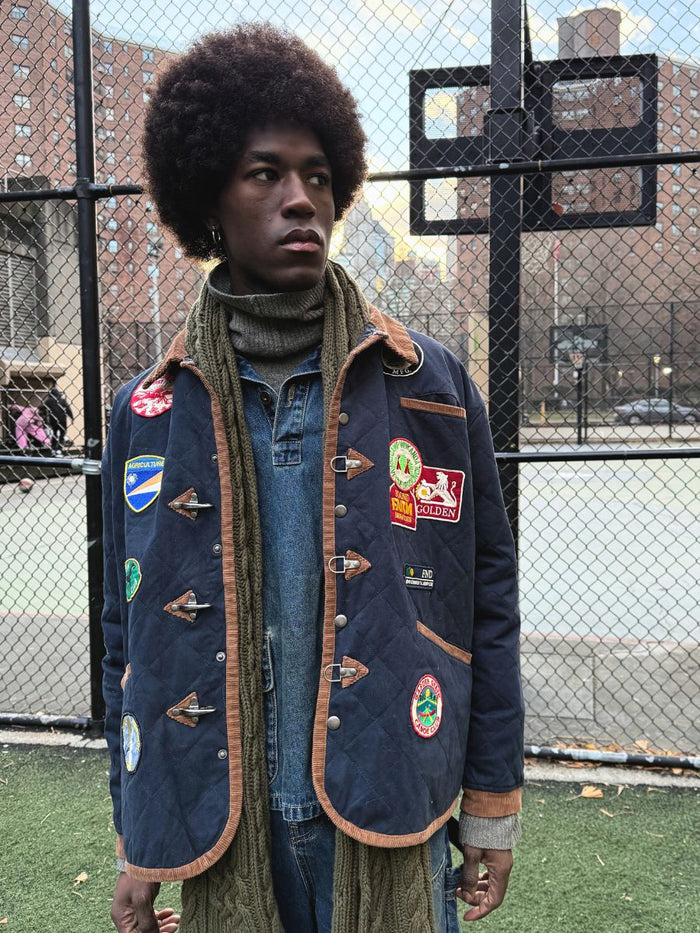 An individual with an afro, wearing a FOUND Farmstead Quilt Patch Jacket and scarf, stands confidently by a sports field fence. The jacket's unisex relaxed fit exudes a cool vintage hunting competition vibe.