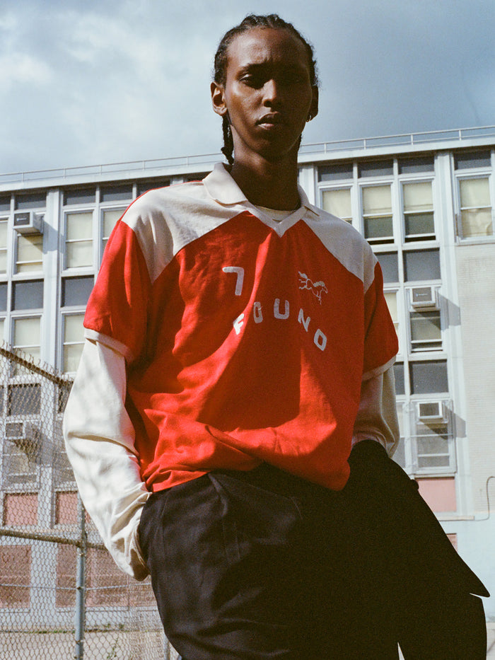 Someone in a Goji Red Jersey by FOUND, featuring the number 7, stands outside near a building with air conditioning units.