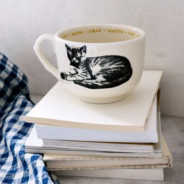 White mug with black cat illustration on a stack of books, blue plaid fabric in the background