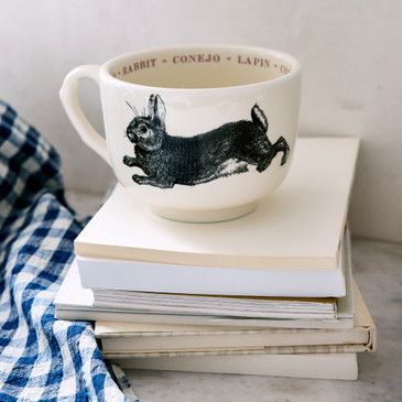White mug with black rabbit illustration on a stack of books with a blue checkered towel.
