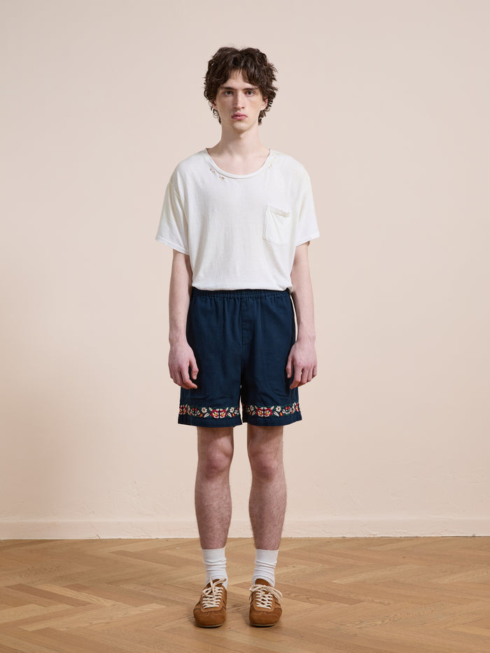 A young person indoors on a wooden floor wears a white t-shirt, Found Horse Equine Twill Shorts with a colorful pattern, white socks, and brown shoes.