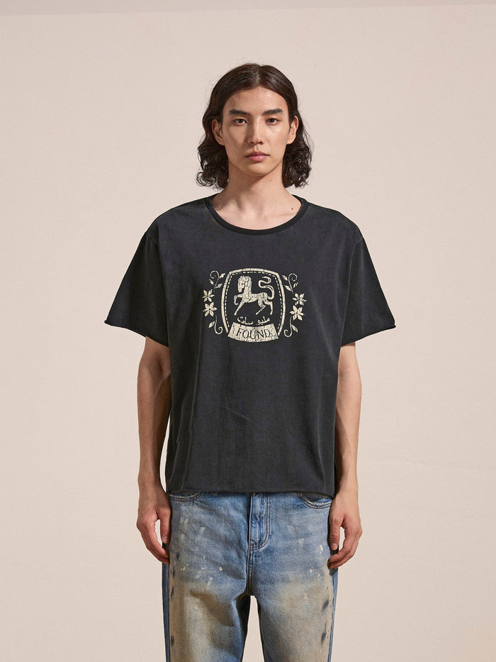A person with long hair, wearing a FOUND Horse Logo Print Tee featuring a crest design, made from 100% cotton and treated with an enzyme wash, paired with light blue jeans, stands against a plain background.