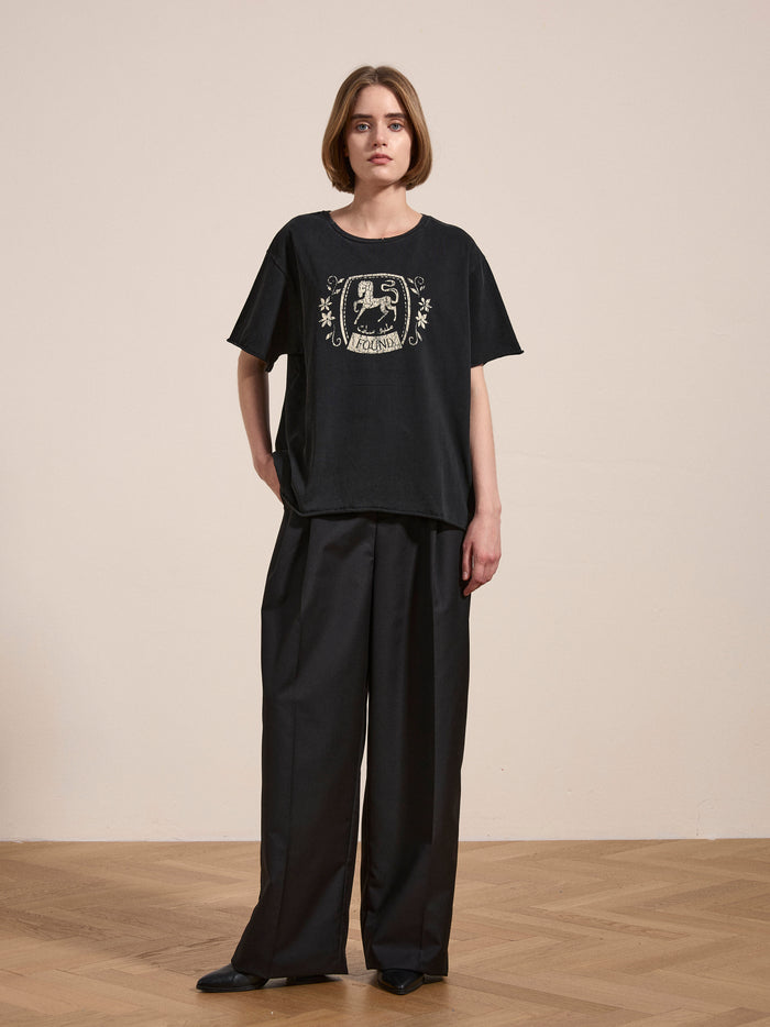 A person stands on a wooden floor wearing a Horse Logo Print Tee by FOUND, made from 100% cotton with an enzyme wash, paired with loose-fitting black pants.