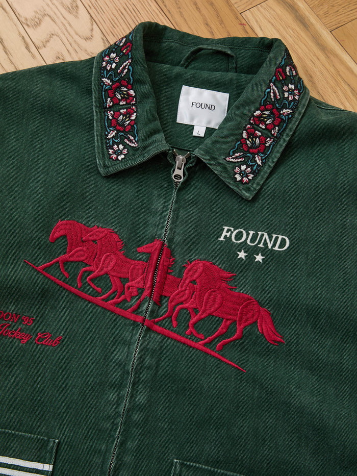 The Found Jockey Club Embroidered Jacket features red racing horses, floral collar details, and "FOUND" with two stars on the chest. This unisex relaxed fit jacket is shown laid flat on a wooden floor.