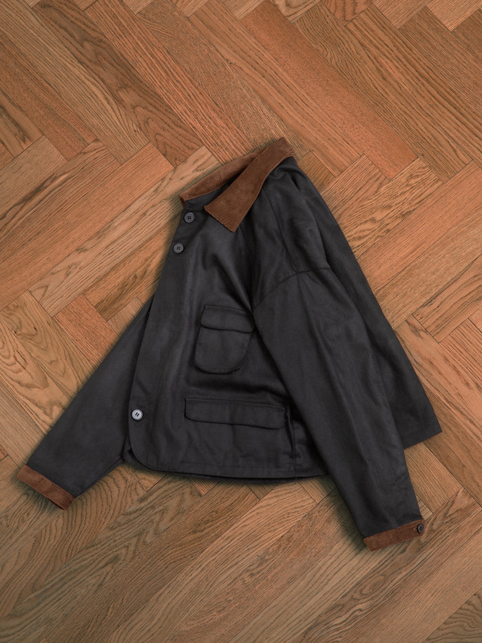 A black Lar Waxed Cotton Box Coat laying on a wooden floor by Found.