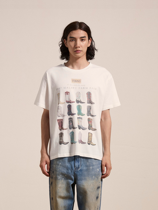 A person with long hair is wearing a Malibu Farm Fair Tee by FOUND, made from soft cotton and adorned with multiple boot images, while standing in distressed jeans against a plain background.
