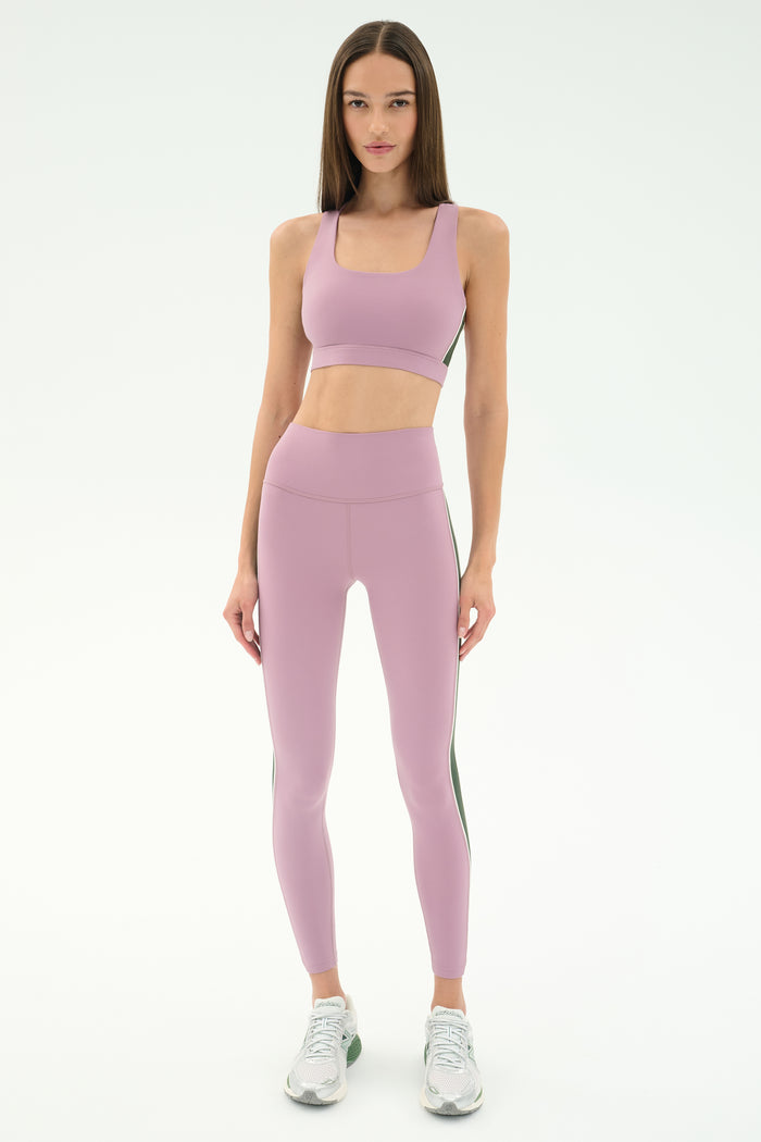 A woman poses in a studio wearing SPLITS59 Margot Rigor Leggings in Blush/Military and a matching light purple sports bra, paired with white athletic shoes.