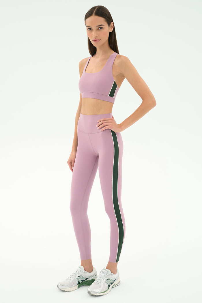A woman stands in a studio wearing SPLITS59's Margot Rigor Legging in Blush/Military, crafted from stretchy nylon-spandex fabric, paired with a matching sports bra and white sneakers.