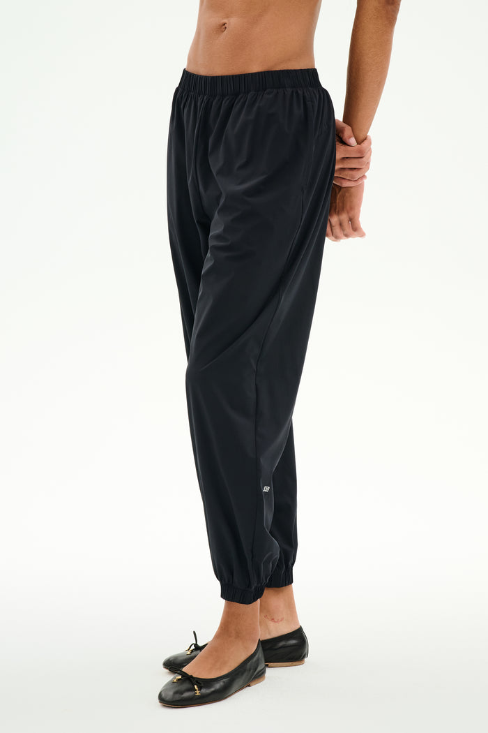 A person wears the SPLITS59 Megan Woven Nylon Jogger in black, featuring a cinched waistband, paired with black ballet flats and standing against a plain white background.