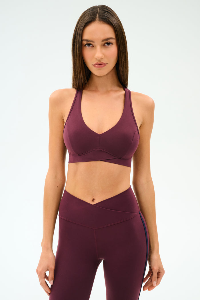 Woman stands against a plain background, wearing the SPLITS59 Mia Rigor Bra in Dark Cherry paired with matching high-waisted leggings.
