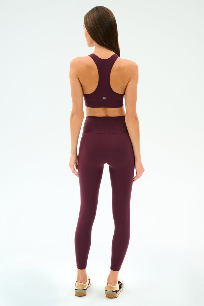 A woman faces away, wearing the SPLITS59 Mia Rigor Bra in Dark Cherry with matching leggings and light sneakers, set against a plain white background.