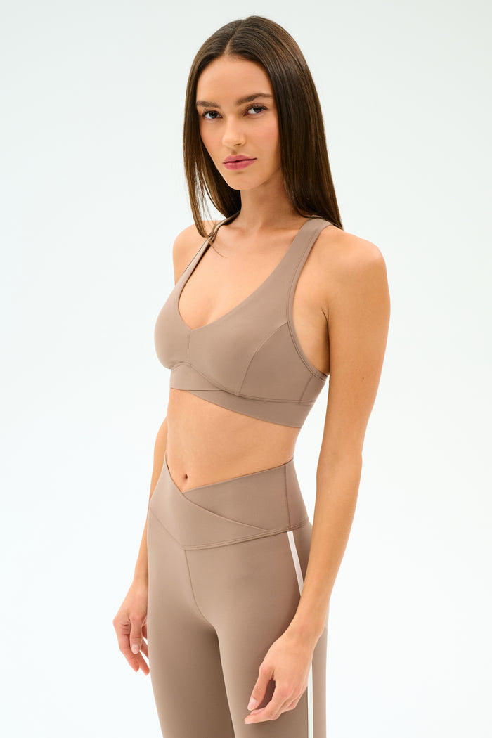 A woman with long straight hair wears the SPLITS59 Mia Rigor Bra in Latte and matching high-waisted leggings, both made from stretchy nylon spandex, against a plain white background.