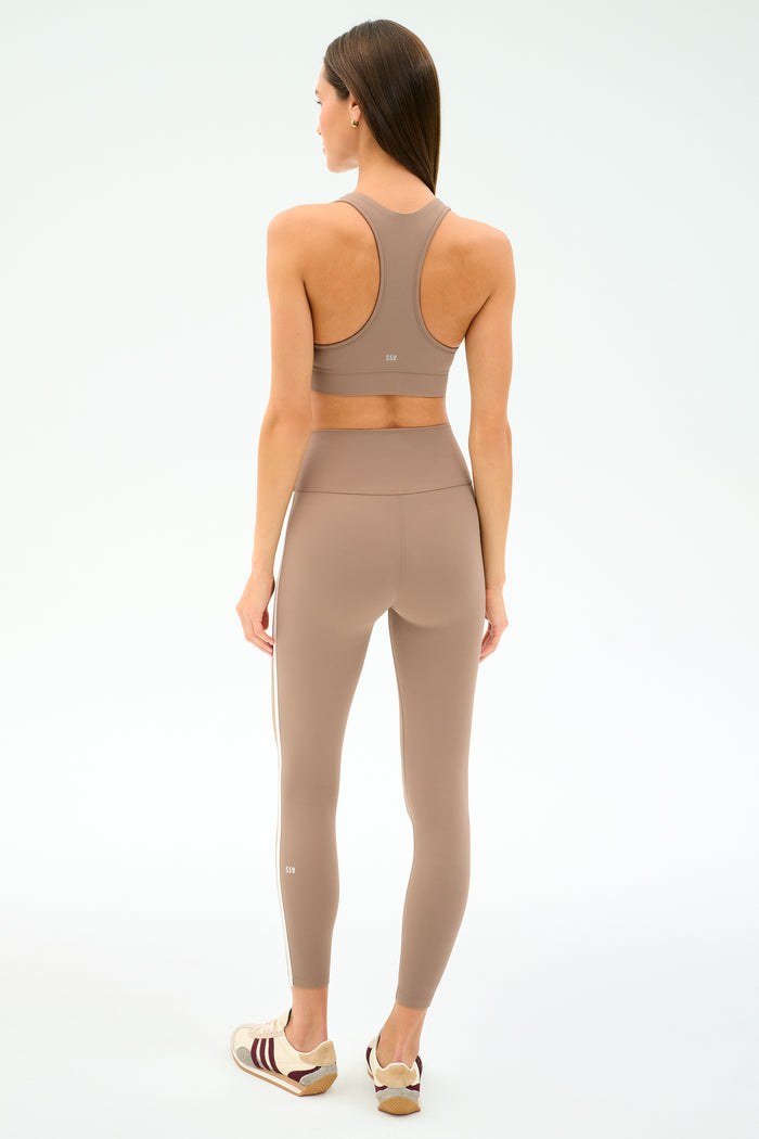 A woman faces away, wearing the SPLITS59 Mia Rigor Bra in Latte with matching leggings and white athletic shoes, set against a plain light background.